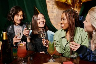 young birthday girl in party hat holding champagne near multiethnic girlfriends and cocktails in bar